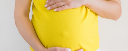 Woman in yellow clothes touching big belly with hand. Isolated on gray background. Emotional loving pregnancy time - 33 weeks. Baby expectation. Happiness and safety concept. Front view. Closeup.