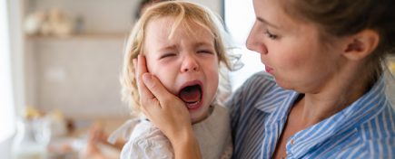 A mother holding a crying toddler daughter indoors in kitchen.